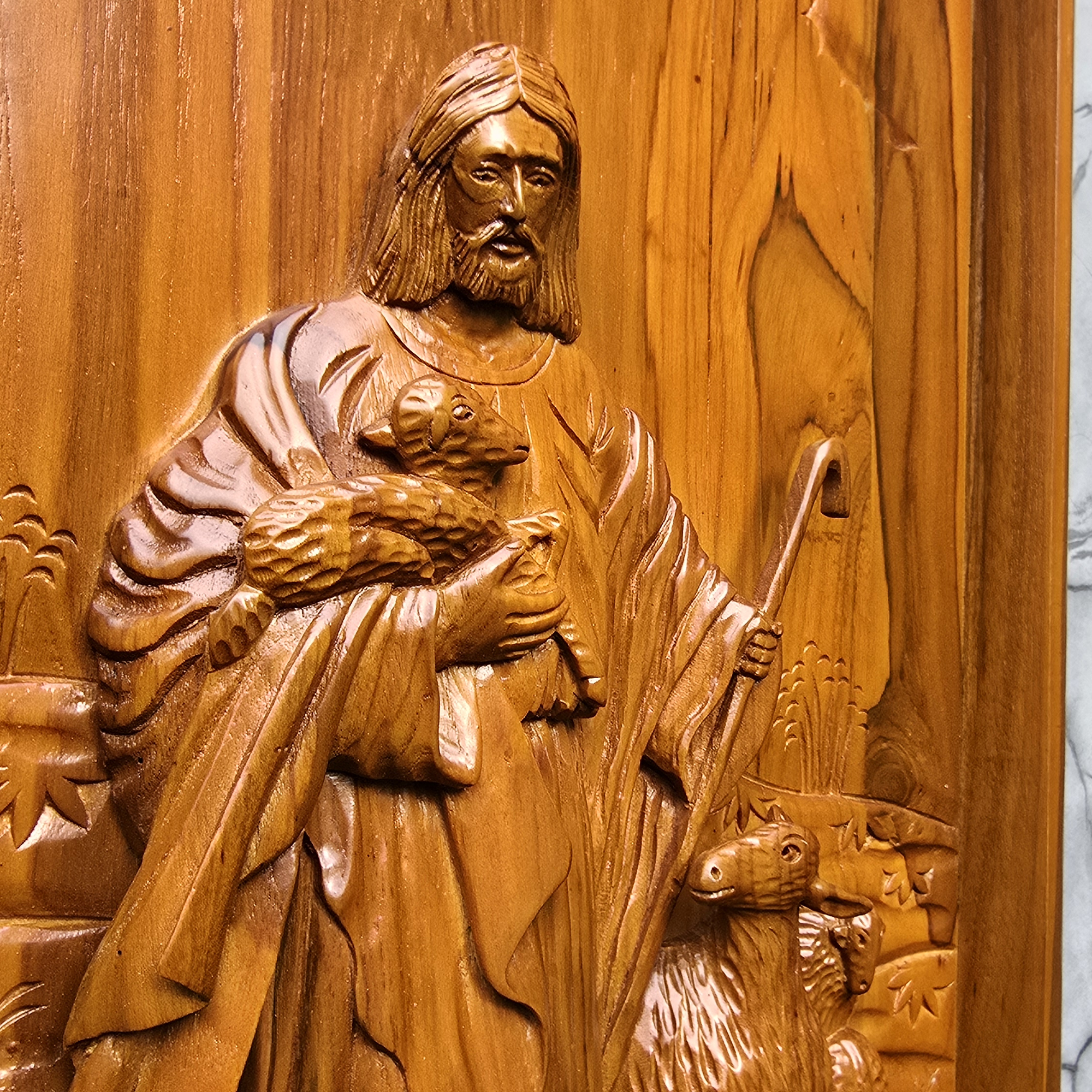 Wooden carving of a shepherd holding a lamb and a staff on a wooden panel.