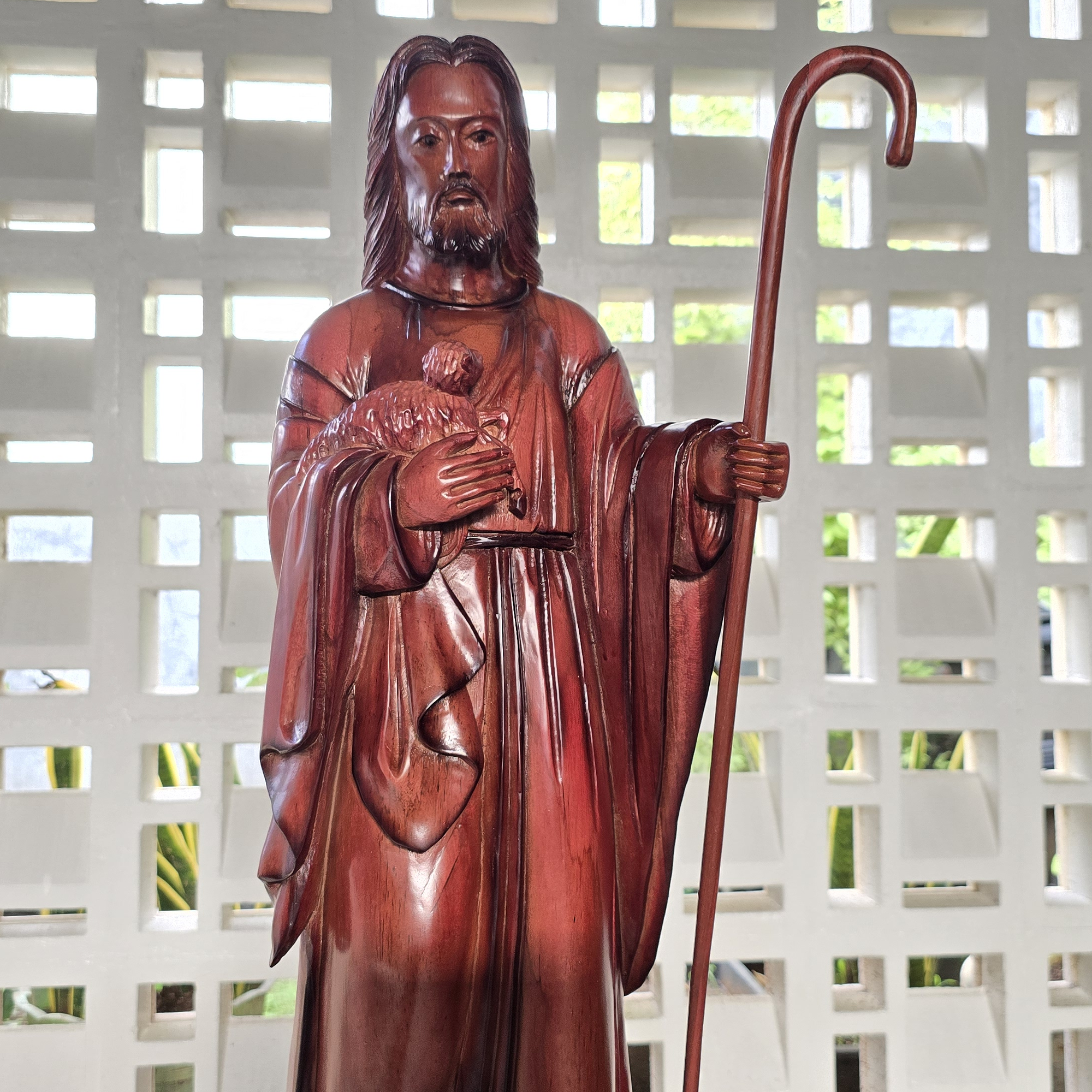 Wooden statue of Jesus shepherd holding a staff and a heart, against a white lattice background.