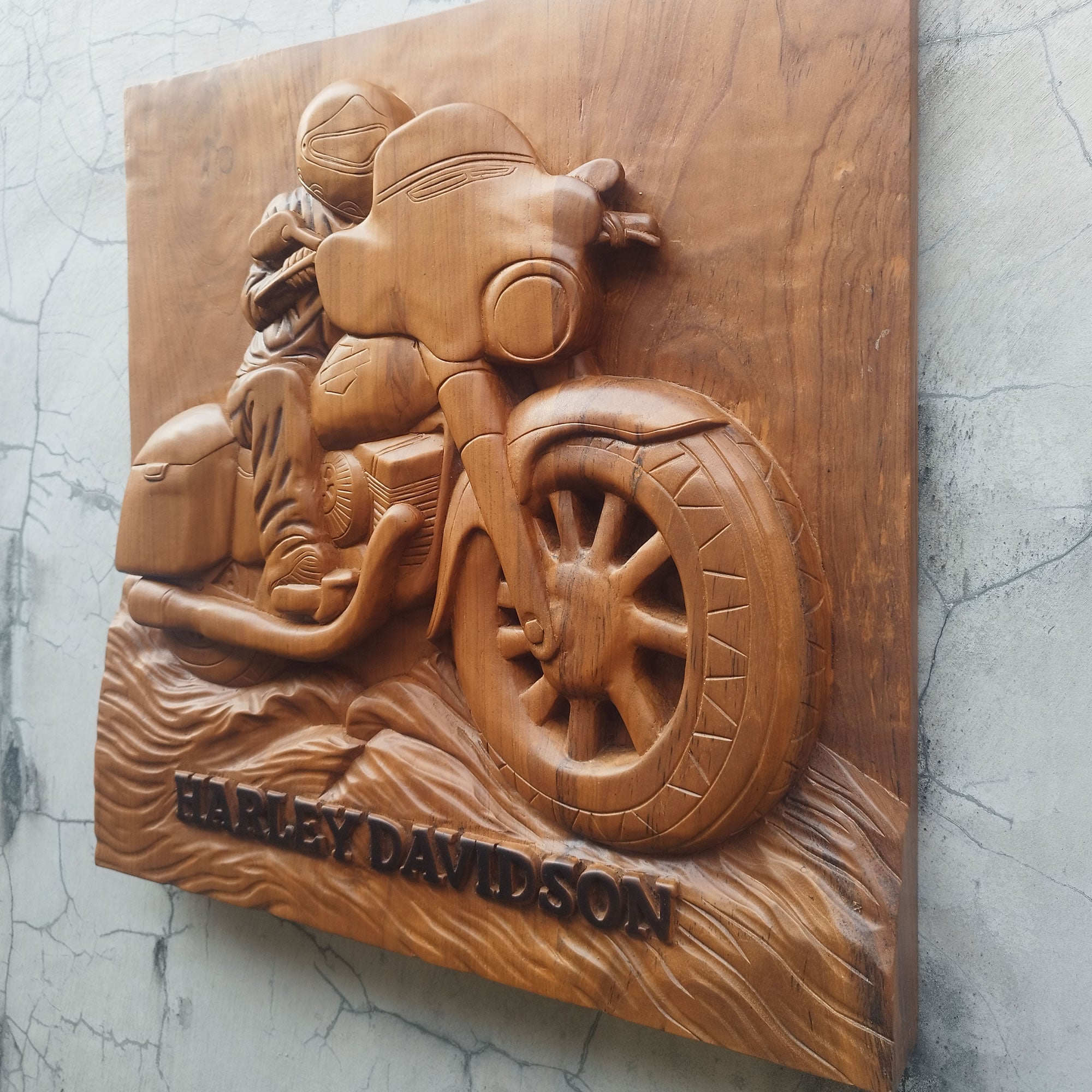 A hand-carved wooden wall plaque featuring a Harley-Davidson motorcycle and rider, with the text 'HARLEY DAVIDSON' carved at the bottom, displayed against a textured gray wall.