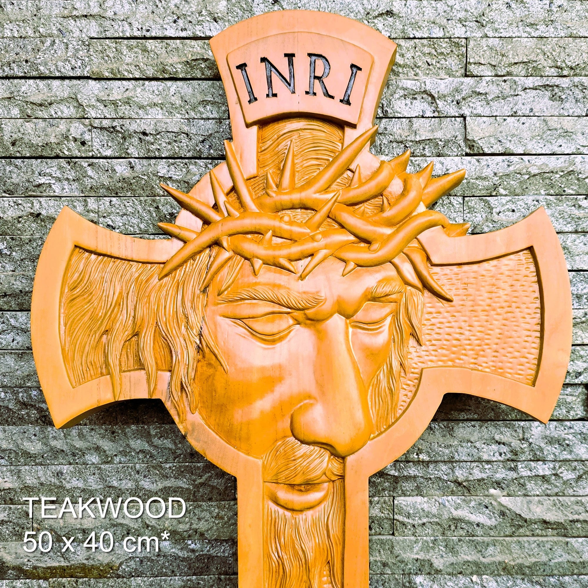 A carved wooden wall cross featuring a face with a crown of thorns and the letters INRI at the top, shown against a gray stone wall. White text in the lower left reads 'TEAKWOOD 50 x 40 cm*'.