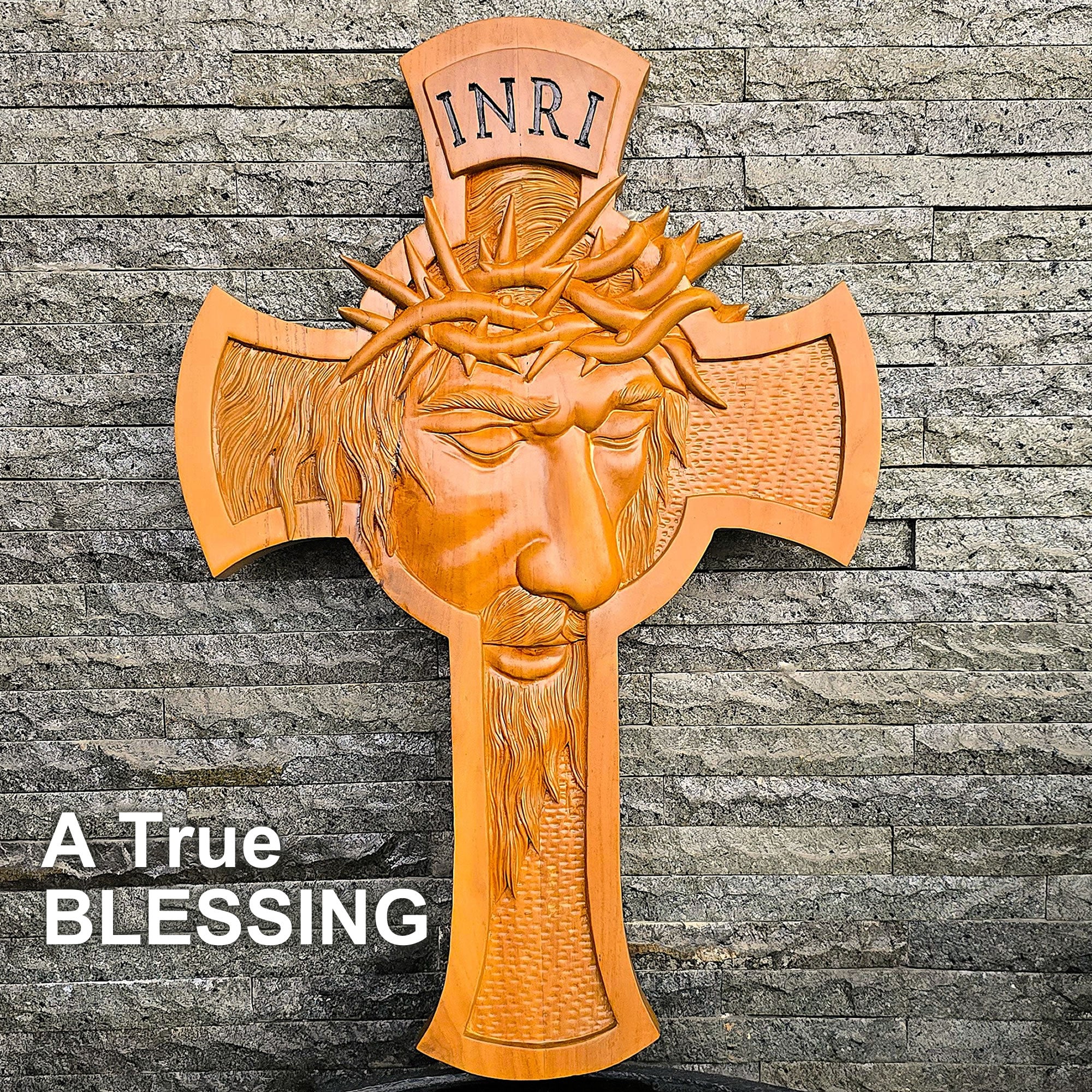 A carved wooden Jesus Christ cross with a crown of thorns and the letters INRI at the top, shown against a gray stone wall. White text in the lower left reads 'A TRUE BLESSING'.
