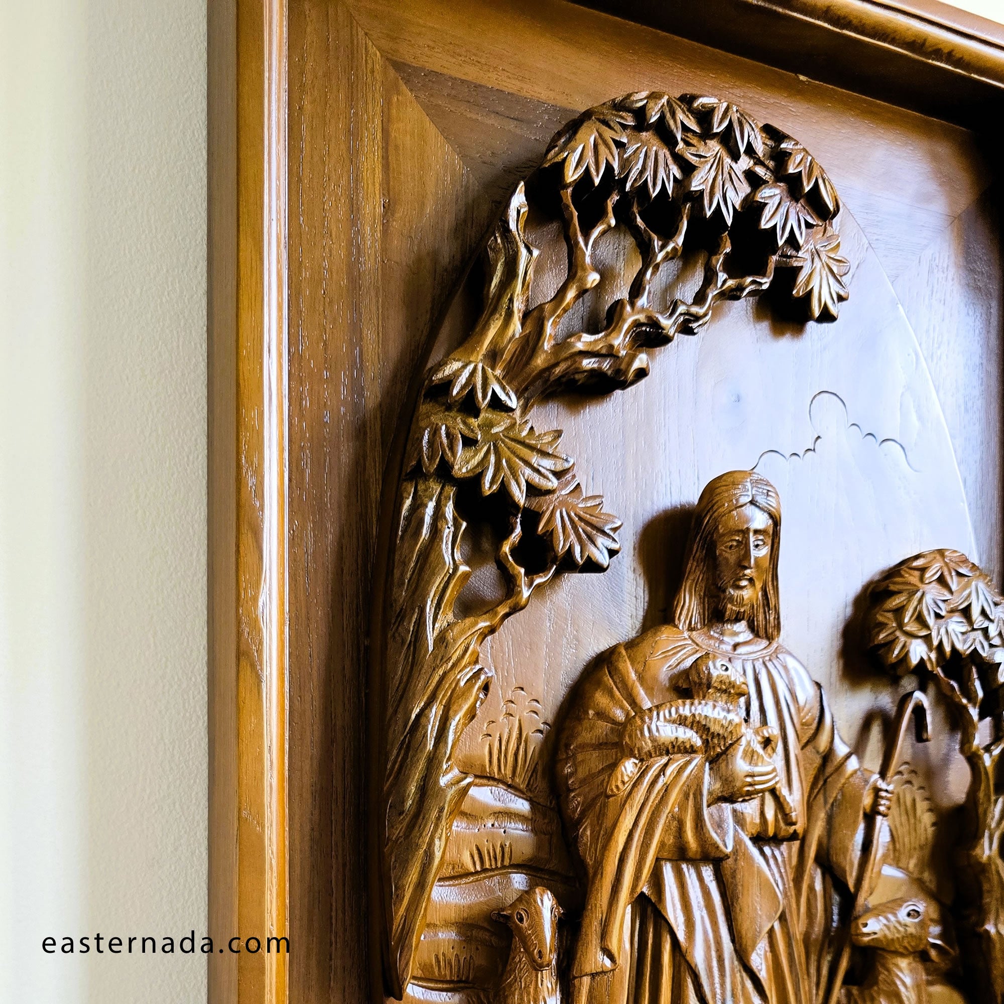 Close-up of a carved wooden religious wall piece showing Jesus Shepard holding a sheep, with leafy tree branches overhead and a warm wood frame visible on the left.