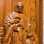 Wooden carving of a shepherd holding a lamb and a staff on a wooden panel.