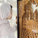 A woman wearing a light pink hijab and white lace top looks at a large, intricately carved wooden wall art piece depicting an Islamic Muslim Hajj Kaaba Teakwood Hand-Carved Sculpture Wall Art Interior Display 