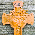 A carved wooden wall cross featuring a face with a crown of thorns and the letters INRI at the top, shown against a gray stone wall. White text in the lower left reads 'TEAKWOOD 50 x 40 cm*'.