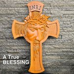 A carved wooden Jesus Christ cross with a crown of thorns and the letters INRI at the top, shown against a gray stone wall. White text in the lower left reads 'A TRUE BLESSING'.