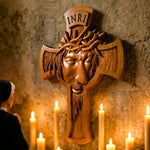 A carved wooden religious cross featuring a face with a crown of thorns and the words INRI at the top, mounted on a stone wall with lit candles in the foreground and a person partially visible on the left.