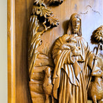 Close-up of a carved wooden religious wall piece showing Jesus holding a lamb, with sheep and leafy branches around him, photographed against a wood-toned background.