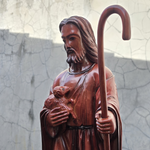 Statue of Jesus the shepherd holding a lamb and a staff against a textured wall.
